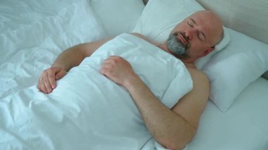 Sabahları uyuyan orta yaşlı sakallı bir adam. Dinlenen erkek yatakta yan yana dönüyor. Yatak odasında yatarken battaniyeyi düzeltiyorum. Uyuyan yetişkin beyaz yatağın altında dinleniyor.