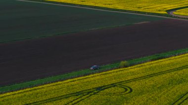 Çiftçi çimenli patika boyunca tarlada tarla sürerken kolza tohumu ve tarla sürüyordu. Kırsal ilkbahar manzarasında, çiçek açan ekinler ve ekilmiş toprak arasında yolculuk yapan bir araç. Parlak sınırlarla çevrili kırsal bölgede hareket eden bir pikap.