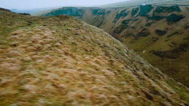 Çimenli yamaçlarla dik dağ vadisinden geçen bir araba. Beyaz araç İngiltere 'nin Peak District bölgesinde dolambaçlı yol boyunca ilerliyor. Engebeli yamaçtan geçen kıvrımlı yolun havadan görünüşü. Resimli yeşil arazide manzaralı sürüş