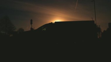 Sessiz yolda yükselen güneşe karşı giden bir kamyon. Sabahın köründe karanlık trafik ve otomobil gölgesi. Büyük araç uzun bir siluet oluştururken altın sabah ışığı yansıtıcı asfalt üzerine yayılıyor.