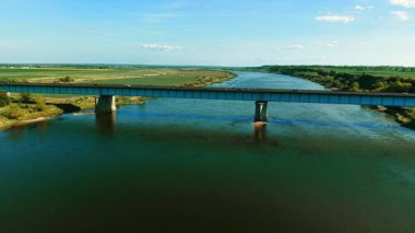 Kırsal alanda geniş nehir üzerinde uzun köprüden geçen arabalar. Araçlar mavi gökyüzünün altında yükseltilmiş yol boyunca ilerliyorlar. Kırsal kesimde akan nehrin yukarısındaki viyadük üzerinde trafik var.