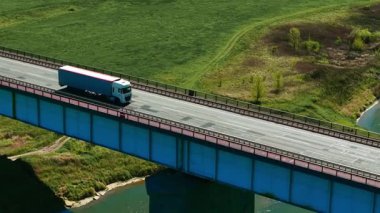 Kırsal bölgedeki köprüden geçen uzun yol tırı. Yük taşıma aracı kargoyu manzaralı kırlara taşıyor. Yükseltilmiş yolda ilerleyen lojistik kamyonu geniş su yolu boyunca ilerliyor.