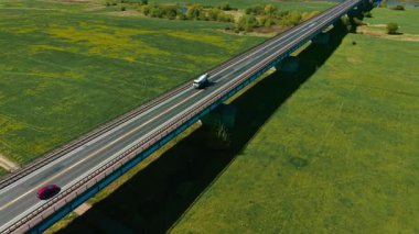 Beton karıştırıcı kamyon ve kırmızı araba yeşil tarlaların üzerinden geçiyor. Çimento kamyonu ve yolcu aracı kırsal alanda yükseltilmiş yol boyunca ilerliyorlar. Karıştırıcı kamyonu ve küçük otomobil kırsal arazide viyadük üzerinde seyahat ediyor.