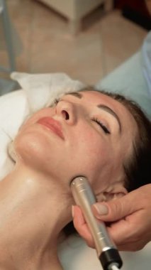 Donanım Yaşlanma Karşıtı Tedavi çene hattı üzerinde çalışıyor, dikey yönelim. Cilt uzmanı, genç görünmek için kadın müşterinin alt yüzüne cihaz temelli masaj yapıyor. Güzellik terapisti cildini tazelemek için estetik aletler kullanıyor.