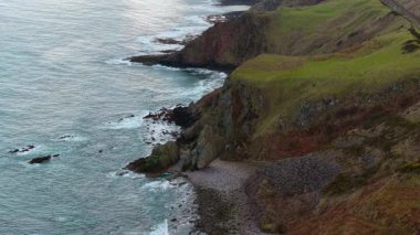 Dalgaların sarp kayalıklara ve kayalık kıyılara çarptığı fırtınalı bir sahil. Vahşi okyanus sörfü bulutlu gökyüzünün altındaki engebeli kıyı şeridine vuruyor. Çimen ve taşla kaplı pürüzlü deniz buluşması. Dalgalı sular yükselmiş kıyı arazisine çarpıyor