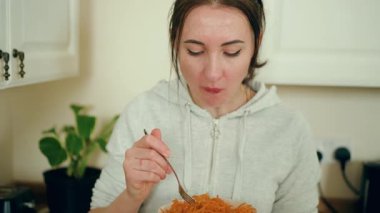 Modern beyaz mutfakta dilimlenmiş havuç tadı alan bir kadın. Çağdaş yemek alanında taze hazırlanmış sebze salatasının tadını çıkaran bir kadın. Bayan şık mutfak alanında besleyici yemeklerden hoşlanıyor.