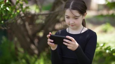 Parkta akıllı telefondan oyun oynayan tatlı bir genç kız. Sevimli ergen kız cep telefonuyla dışarıda eğleniyor. Mobil kablosuz teknolojiler, yenilikler, kusursuz bağlantı ve iletişim