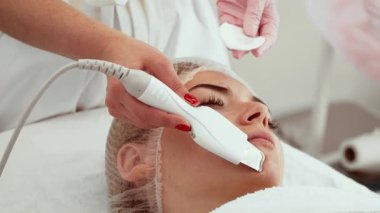 Güzellik uzmanı kaplıca salonunda deri dökülmesi için ultrasonik cilt fırçası kullanıyor. Uzman, modern aletle nazikçe temizlik tedavisi uyguluyor. Cilt tazeleme ve hidrasyon tedavisi gören bir müşteri.