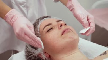 Kadın hasta kozmetoloji kliniğinde dinleniyor, güzellik uzmanı nazikçe cildini temizliyor. Doktor, güzellik prosedürünü titizlikle gerçekleştiriyor. Cilt bakımı uzmanı ferahlatıcı ve sakinleştirici tedaviye odaklanır