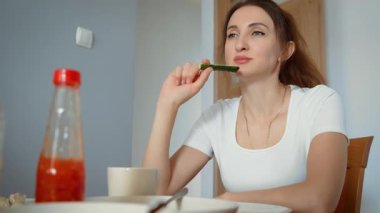 Yemek sırasında salatalık yiyen genç bir kadın. Taze sebzelerden hoşlanan düşünceli bir kadın. Hanımefendinin sakin bir yemek anında sağlıklı salatalıkların tadını çıkarması.