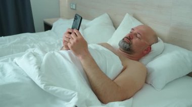 Akıllı telefon tutan, sabah yatakta yatarken sosyal medyayı kontrol eden bir adam. Uyandıktan sonra battaniyenin altında mobil uygulamalara bakmak. Konforlu yatak odasındaki güncellemeler için telefon kullanmak. Erken saatlerde cihazla yatakta dinlenmek