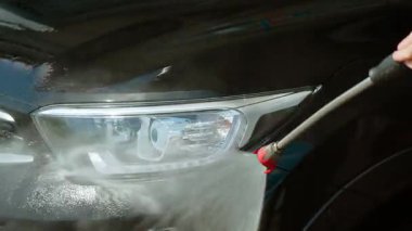 Otomatik yıkamada yüksek basınçlı suyla araba farlarını durulayan tanınmaz halde bir kadın. Güçlü su jeti kullanan bir kadın arabanın ön ışığını püskürtüyor. Basınçlı akış ile far bölgesini temizleyen kişi