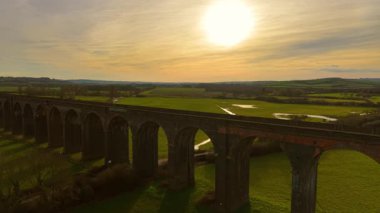 Demiryolu viyadük alevli günbatımı gökyüzünün altında dimdik duruyor. İnsansız hava aracından görülen eski bir viyadük. Resmedilmeye değer bir manzaraya yerleştirilmiş. Tarihi yerler, ünlü alanlar, nefes kesici doğa yoluyla altyapı.