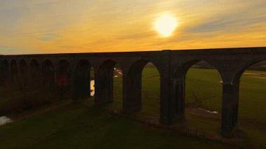 Gün batımında kırsal alana yayılan görkemli demiryolu viyadüğü. Alacakaranlıkta manzaralı kırsal bölgeye uzanan yükseltilmiş viyadük kanalı. Büyük tuğla köprü, yeşil vadinin üzerinde duruyor, altın akşam gökyüzünün altında.