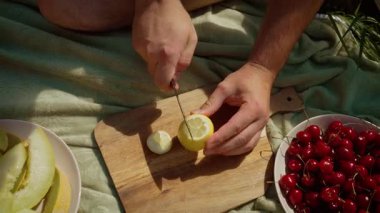 Tahta kesme tahtasıyla limon dilimleyen adam içecekler için taze narenciye hazırlıyor. Erkek eller dikkatlice sarı limonu parçalara ayırıyor. Aile yemeği için meyveleri ustalıkla taşıyan bir adamla açık hava piknik sahnesi.
