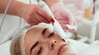 Kozmetik atölyesinde cilt bakımı sırasında ultrasonik temizlik aleti kullanan bir profesyonel. Derin gözenek temizleme ve gençleştirme için gelişmiş bir cihaz uyguluyor. Müşteri dinlenirken profesyonelce nazikçe derisini yüzüyor ve su kaybediyor.