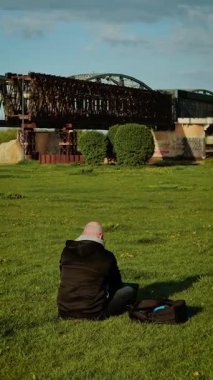 Man sitting on grass working on laptop near railway bridge, vertical orientation. Freelancer focusing on screen in scenic industrial location. Remote worker using computer beside structure in open field. Digital nomad engaging in task with backpack