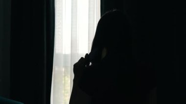 Kadının silueti, pencerenin önünde elinde saç fırçası, yumuşak sabah ışığıyla. Barışçıl otel odasındaki perdenin yanında güne hazırlanan kadın figürü. Kumaşın içinde gün ışığının parladığı yerde sessizce cam kenarına tıkılıp duran kadın.