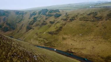 Dik yeşil vadide dolambaçlı yol boyunca giden arabalar. Birleşik Krallık 'ın Peak District bölgesinde asfalt yolda giden araçlar. Engebeli kırsal bölgeyi kesen kıvrımlı yolun havadan görünüşü. Resimli sürüş tepelik arazide çimenlerle