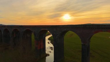 Büyük tuğla viyadük altın gün batımında dolambaçlı nehrin üzerinde yayılıyor. Sıcak günbatımı ışığı tarafından öpülen viyadük manzarası. Parlak gökyüzünün altında manzaralı vadi boyunca uzanan görkemli demiryolu köprüsü