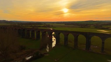 Gün batımında sakin vadide ilerleyen ikonik demiryolu viyadük kanalı. Antik İngiliz demiryolu köprüsü gün batımında parlıyor. Sıcak alacakaranlık gökyüzünün altında engin kırlara uzanan tarihi tuğla yapı.