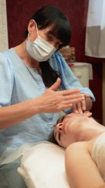 Koruyucu maskeli masaj terapisti alnında ruhani yüz tedavisi uyguluyor, dikey yönelim. Cilt bakımı uzmanı gevşemek için üst yüz kaslarını uyarıyor. Güzellik uzmanı cilt yenilenmesini hızlandırmak için yumuşak hareketler uyguluyor