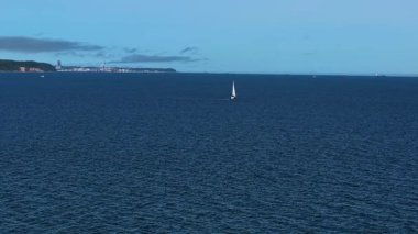 Sakin sularda hareket eden küçük yelkenli yatlı deniz manzarası. Mavi dalgalarda süzülen beyaz yelkenli ile okyanus manzarası. Denizcilik sahnesi açık denizde yalnız bir yelkenli gösteriyor. Su ufku berrak gökyüzünün altında seyir halindeki yatın arkasında uzanıyor.