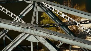 Nehir kıyısındaki demiryolu köprüsünün çelik kirişlerinde oturan güvercinler. Kuşlar, su ve ağaçların yanındaki güneş ışığı altında perçinlenmiş demir yapıda toplanıyorlar. Güvercinler kiriş eklemleri üzerinde dinleniyor ve geometrik desendeki köprünün metal geçitleri