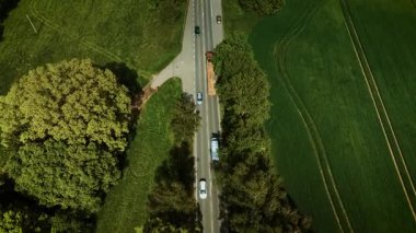 Taşıma aracı kırsal yol boyunca sürüyor. Beyaz hatchback gökyüzü manzarası altında geziniyor. Araba yeşil kırsal alanda hareket ediyor. Araç tarlalar ve ağaçlar arasında açık yolda ilerliyor.