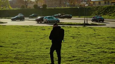 Sırt çantalı bir adam gün batımında Avrupa 'da araba parkında yürüyor. Arka planda araçlar ve binalarla yeşil alanda gezinen bir gezgin. Park halindeki arabalarla caddeye doğru ilerleyen şehir turisti. Sıcak altının tadını çıkaran kişi