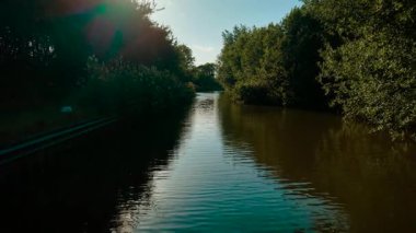 Gölgeli ormanlık alanda kanal kıvrımları. Su, öğleden sonra güneşinin altında parlıyor. Huzurlu İngiliz su yolunu çevreleyen yoğun yeşillik. Sakin sahne, yaz sükunetini yansıtıyor.
