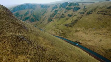 Dik çim vadisinden geçen virajlı yolda giden arabalar. Birleşik Krallık 'ın Peak District bölgesinde asfalt yolda giden araçlar. Engebeli kırsal bölgeyi kesen kıvrımlı yolun havadan görünüşü. Resimli sürüş tepe arazi yeşil ile