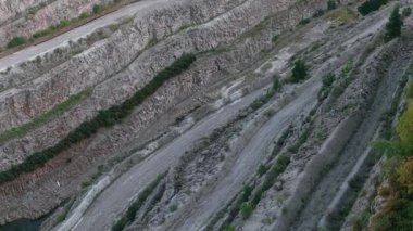 Teraslı taş ocağı duvarları tozlu yollarla ve çıkıntılardan büyüyen seyrek ağaçlarla iniyor. Çürümüş kaya katmanlarının içinden geçen kazı yolları ve yeşil yamalar ortaya çıkıyor. Madencilik eğimleri açıkta kalan katmanlar ve dağınık bitki örtüsü boyunca aşağıya doğru kıvrılıyor.