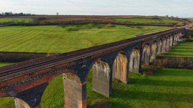 Çimenli yeşil kırlara uzanan tarihi demiryolu viyadük kanalı görüntüsü. Viyadük vadide kavis çiziyor, gün batımında yıkanıyor. Büyük tuğla köprü altın gece parıltısıyla yuvarlanan tarlalar boyunca uzanıyor.
