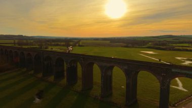 Ilık gün batımı eski viyadük üzerinde gölgeler bırakıyor. Gökyüzünden alınmış yemyeşil vadideki tren köprüsünün gün batımı manzarası. Tarihsel işaretler, ünlü bölge, manzaralı arazide ulaşım yolu.