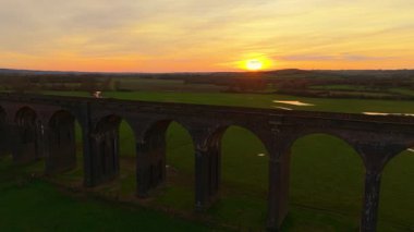 Gün batımında kırsal alana uzanan uzun tuğlalı viyadük. Günbatımı, tarihi viyadük üzerine sıcak gölgeler düşürüyor. Yükselen demiryolu köprüsü parlayan akşam gökyüzünün altında yuvarlanan yeşil araziye yayılıyor.