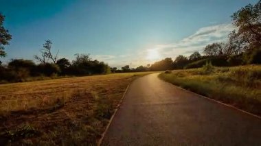 Gün batımında açık alanda virajlı yoldan aşağı iniyoruz POV. Akşamüstü gökyüzünün altında parlayan ufka doğru bisiklet sürmek, bakış açısı. Güneşin aydınlattığı asfalt yolda ilerlerken altın ışık önümüzde, ilk bakışta