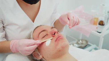 Gözleri kapalı bir kadın, görünürde bir burun halkası var. Kozmetik uzmanı, ahşap aplikatör kullanarak antiseptik krem sürüyor. Güzellik uzmanı yanağına krem sürerken kadın cilt bakımı sırasında dinleniyor. Rahatlatıcı bayan...