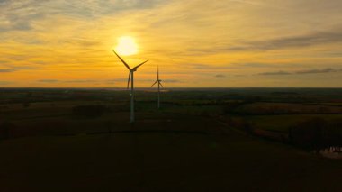 Yel değirmenleri gün batımında yeşil enerji üretiyor. Rüzgâr türbini dramatik turuncu gökyüzü altında temiz enerji sağlamak için uzun bıçaklar kullanıyor. Rüzgâr pervaneleri rüzgar çiftliğinde temiz sistemlere güç veriyor.