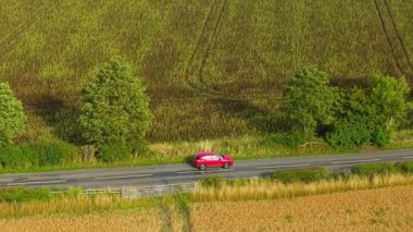 Sıcak akşam ışığında kırsal yolda kırmızı bir SUV sürüyordu. Gün batımında yeşil çitleri ve altın tarlaları geçen parlak bir geçiş. Turuncu gökyüzünün altında tarla yakınlarında seyahat eden kırmızı spor aracı