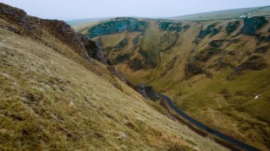 Araba dik çim vadisinden geçen virajlı yolda ilerliyor. Araç İngiltere 'nin Peak District bölgesinde manzaralı bir rotada ilerliyor. Etrafı engebeli yamaçlarla çevrili asfalt yolda dolaşan kırmızı araba. Tepenin içinden geçen resimsel sürüşün havadan görüntüsü