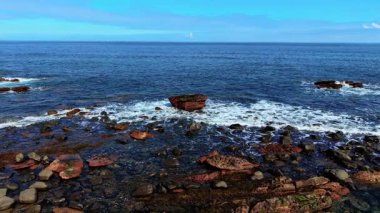 Dalgalar kıyı kayalarıyla çevrili büyük kırmızı kayalara çarpıyor. Dağılmış taşların ve dalgalı levhaların üzerinde dönen deniz. Okyanus engebeli kıyı boyunca yıpranmış oluşumlara çarpıyor.