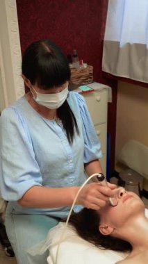 Darsonval Cilt Uyarımı yanaklarda, dikey yönelimde. Güzellik terapisti yüksek frekanslı elektrot masajı uyguluyor. Tirajını arttırmak için kadın müşterinin yüz derisine. Deriyi geliştirmek için Darsonval aygıtı kullanan Cilt Uzmanı