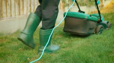 Elektrikli çim biçme makinesinin arkasında lastik çizmelerle yeşil çimlerin üzerinde yürüyen ayaklar. Wellingtons 'da kordonlu kesiciyle çimleri biçerken bacaklar arka bahçede hareket ediyor. Süsleme sırasında elektrikli biçme makinesi kullanırken çimenlere basan botlar
