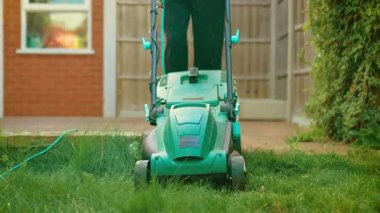 Ön taraf elektrikli çim biçme makinesine bakıyor. Bahçede uzun otlar kesiyor. Tuğla evin yakınındaki çim biçme makinesini iten kişi. Kalın bitki örtüsünde kordonlu çim biçme makinesi kullanılıyor.