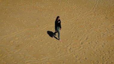 Çıplak ayakla yürüyen bir kadın dağınık ayak izleriyle boş kumlu arazide. Güneşli sahil yüzeyinde tek başına ilerleyen bir kadın. Altın kumda sakince gezinen kadın arkasında ışık izi bırakıyor.