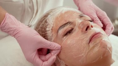 Güzellik kliniğinde yüz hidrasyon maskesi uygulanan bir kadın. Tedavi sırasında şeffaf maskeyi dikkatlice yerleştirmek. Klinik ayarlarda nemlendirici cilt oturumu sırasında istemci dinleniyor