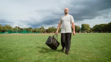 Büyük bir çanta taşıyan bir adam parkta yürüyor. Yetişkin adam düşünceli ve sakin görünüyor. Orta yaşlı, açık tişörtlü, siyah pantolonlu ve sağlam ayakkabılı bir adam. Sakallı adam çimenli alanda geziniyor geniş bir çanta tutuyor.