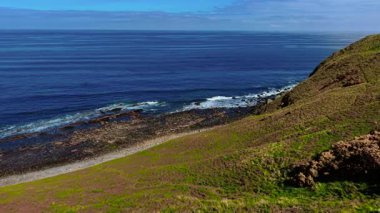 Okyanus dalgaları çimenli uçurumun dibindeki kayalık kıyıya vuruyor. Kıyı şeridi boyunca yavaşça dalgalanan dalgalar. Yeşil yamaçların kayalara dönüştüğü deniz kıyısı. Kıyıdaki su, engebeli arazinin yakınındaki çakıllı plaja doğru akıyor.