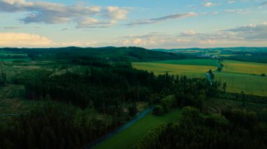 Sakin bir akşam gökyüzü altında uzanan açık kırsal bölge. Yeşil çayırların etrafını saran yuvarlanan tepeler. Dağınık bulutların altında uzanan huzur dolu bir manzara.